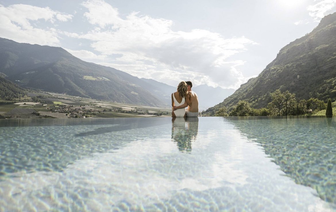 Ein Mann und eine Frau sitzen am Rand eines Pools mit Landschaftskulisse im Hintergrund